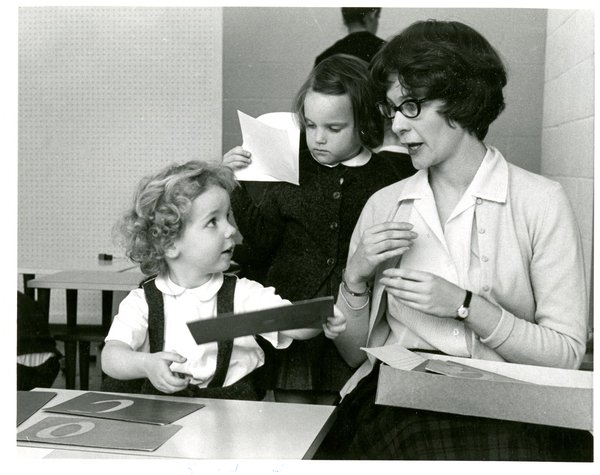 Photograph of a child using sandpaper letter while looking at teacher. Other students work in the background.