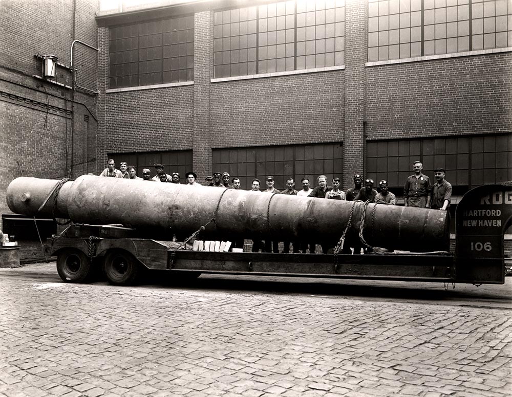 Photo from Archives & Special Collections Business Collections, line of men standing behind a large beam on a truck bed in front of a factory-like building.